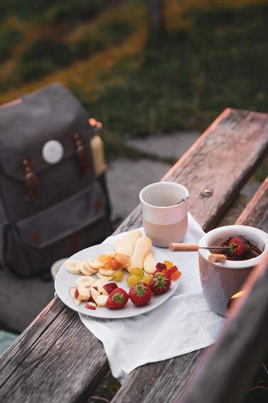 A picnic setup with a plate of fruit, a mug, and a chocolate dessert on a wooden bench. 2