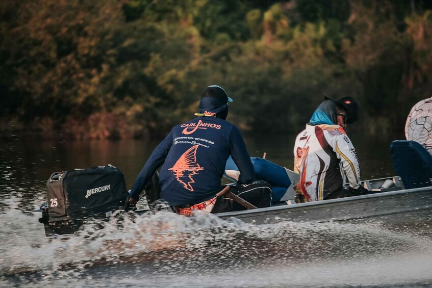 A person in a boat wearing a jacket with a winged logo, a bag labeled 'MERCURY,' and another person in a patterned jacket. 1