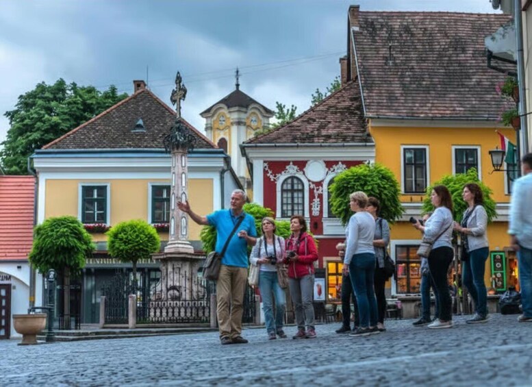 A cobblestone street with historic buildings, a central fountain, and a group of people standing and conversing. 1
