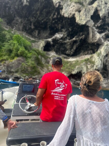 A person in a red shirt with 'Captain Dio' on the back, steering a boat with a rocky shoreline in the background. 4