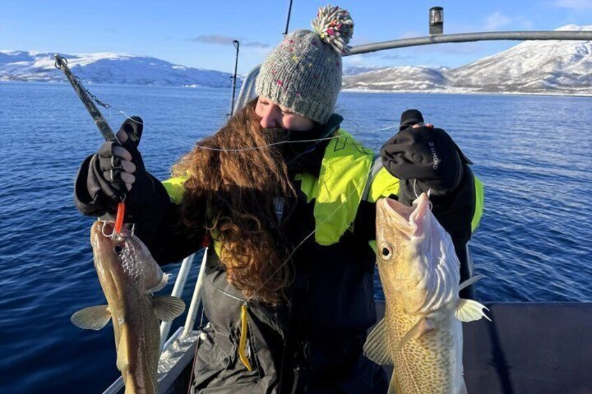 A person holding two fish, wearing a bright yellow jacket, on a boat with a snowy landscape in the background. 3