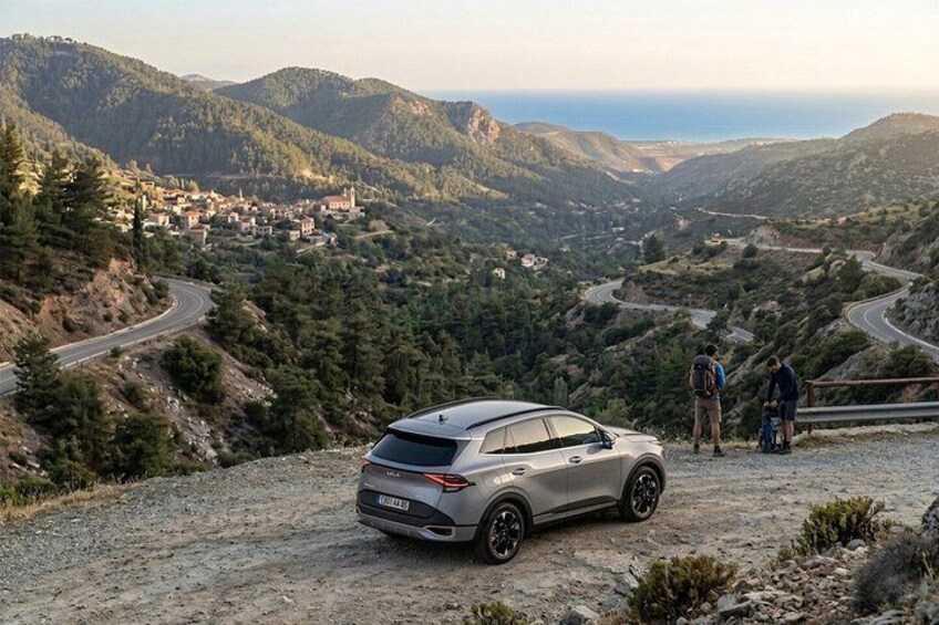 A silver SUV parked on a dirt road with a scenic view of a mountainous landscape and a village in the distance. 2