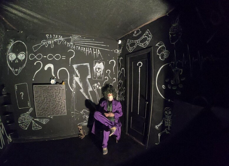A person sitting in a room with graffiti, including a maze, skulls, and a jester mask. 4