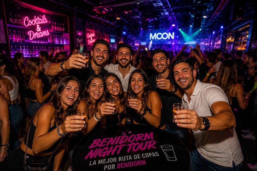 A group of people at a bar with neon signs and a round table labeled 'BENIDORM NIGHT TOUR.' 1
