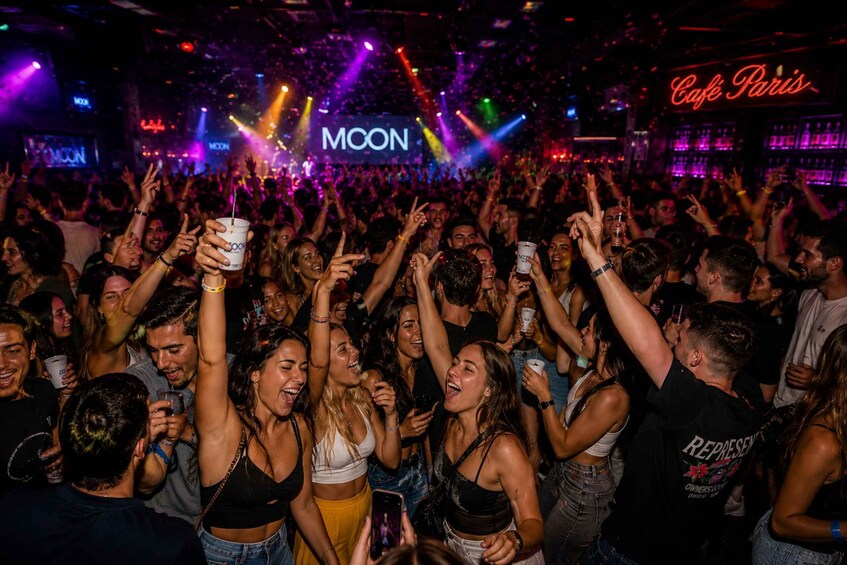 A crowded nightclub with people dancing and holding drinks, with a neon sign reading 'Cafe Paris' and a stage with 'MOON' lighting. 2