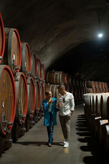 A person in a blue coat and another in a white shirt walking through a wine cellar with rows of wooden barrels. 1
