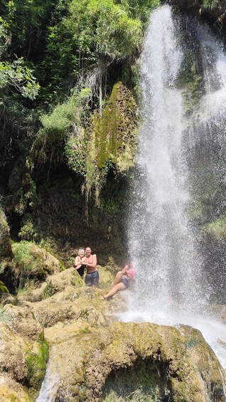 A waterfall in a forested area with people relaxing on rocks. 4