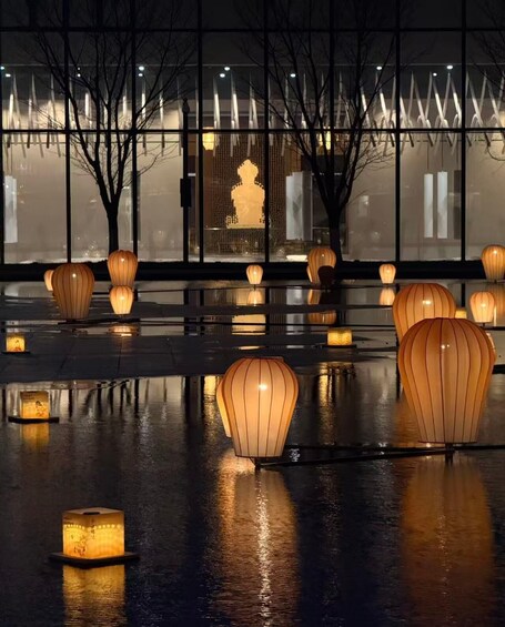 A series of illuminated paper lanterns on a wet reflective surface, with a silhouette of a person in the background through a window. 4