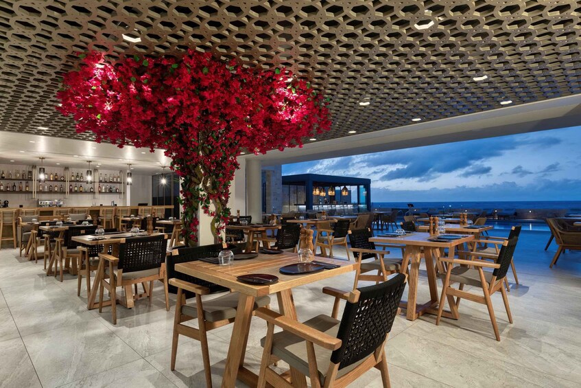 A restaurant with wooden tables and chairs, a large artificial red flower arrangement, and a view of the ocean. 4