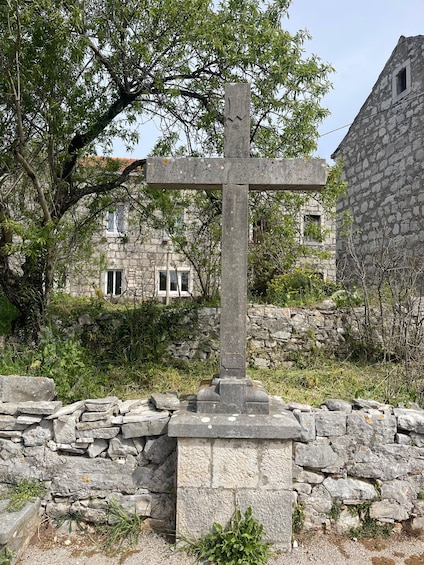 A stone cross with inscriptions, surrounded by a stone wall and greenery, with a building in the background. 3