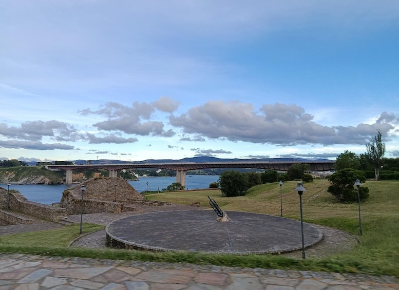 A circular stone stage with a sculpture in the center, a stone wall, a bridge, and a body of water in the background. 1
