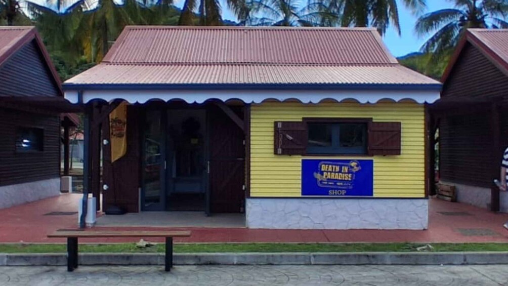 A small shop with a yellow facade and a blue sign that reads 'DEATH IN PARADISE SHOP.' 5