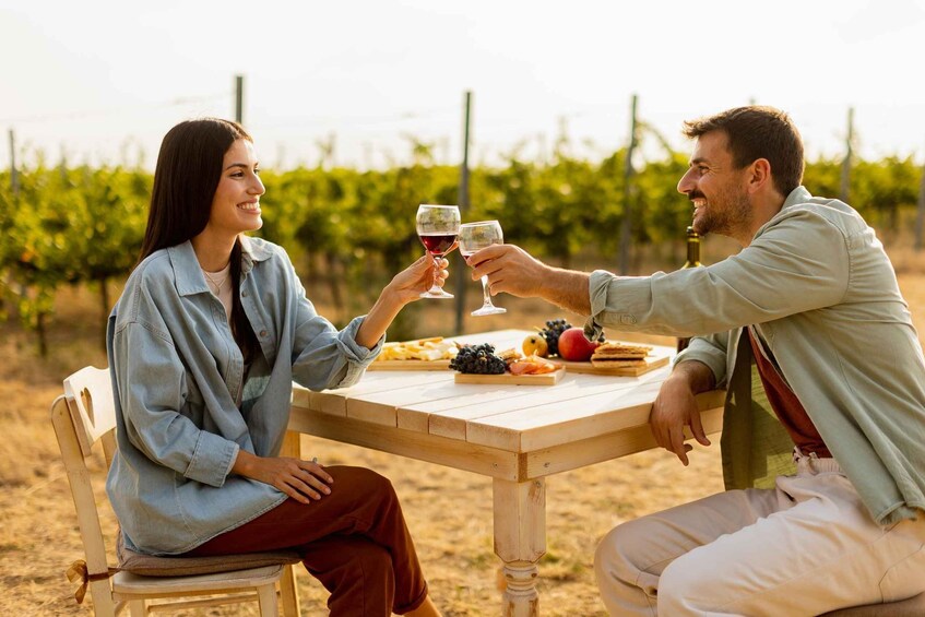 Two people toasting with wine glasses at a wooden table with a vineyard in the background. 1