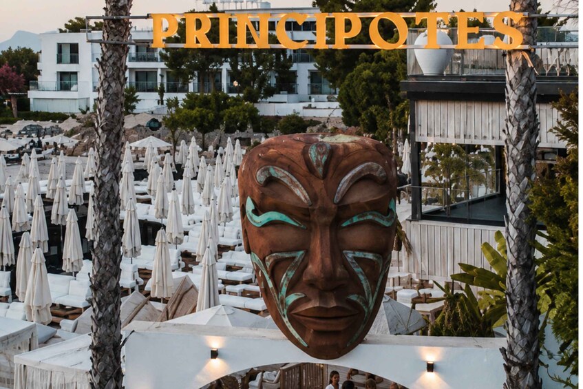 A large bronze mask sculpture in an outdoor setting with white lounge chairs and umbrellas. 2