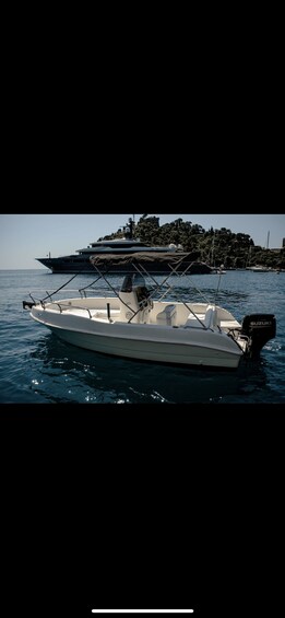 A white motorboat with a Suzuki engine on calm waters, a larger yacht in the background, and a hilly landscape with trees and buildings. 2