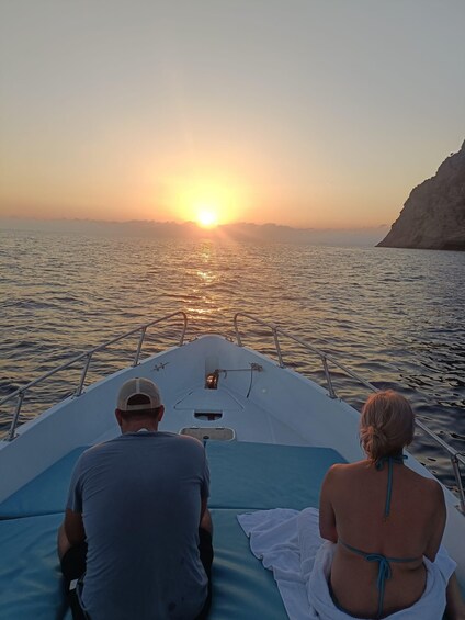 A boat with two people facing the sunset over calm waters. 1