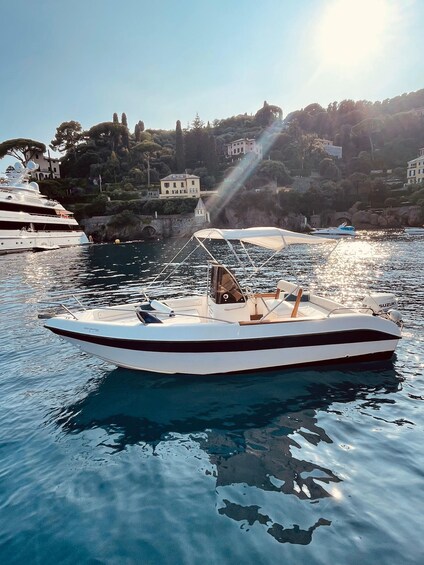 A white speedboat with a Suzuki engine on calm water, a large yacht docked nearby, and a coastal town with buildings and trees in the background. 3