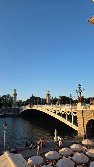 A bridge with ornate statues and lamp posts, a river below, and people sitting at outdoor tables with umbrellas. 3