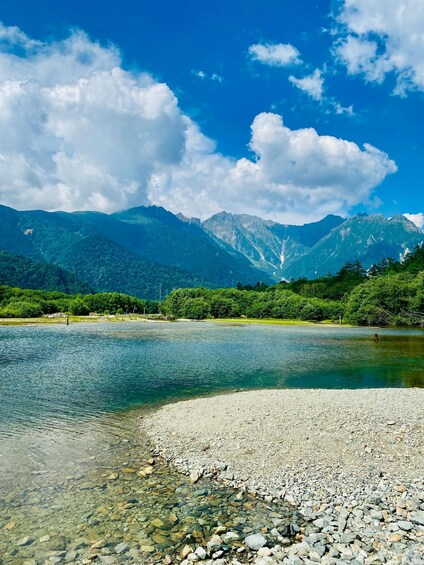 A clear lake with a rocky shore, surrounded by lush green mountains and a blue sky with clouds. 5