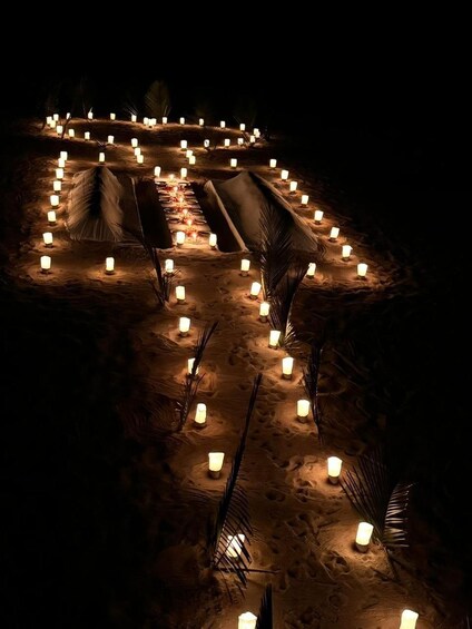 A nighttime outdoor setting with a series of tents and candles arranged in a circular pattern on sandy ground. 1