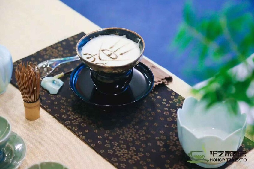 A traditional tea ceremony setting with a black tea bowl on a patterned tray, a whisk, and a white tea cup. 1