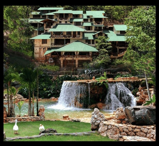 A resort with multiple green-roofed buildings, a waterfall, and a pond with ducks. 2