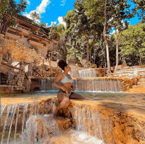 A person sitting on a rock by a cascading waterfall in a natural setting with trees and stone structures. 1
