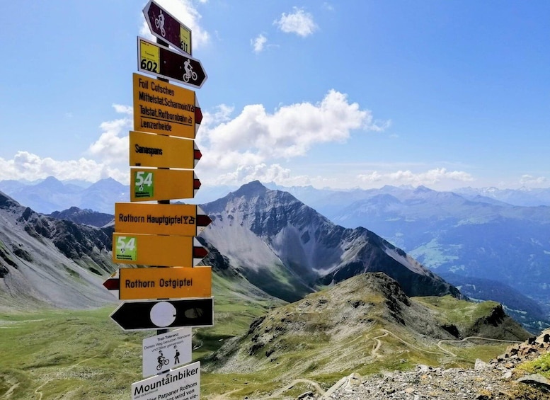 A mountain trail signpost with multiple directional arrows, set against a backdrop of a mountainous landscape. 1