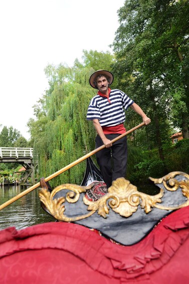 A gondolier in a striped shirt and hat, standing in a gondola with a wooden oar, in a canal with willow trees and a bridge in the background. 5