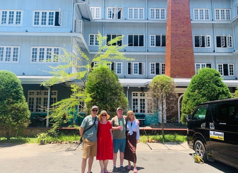 A group of people standing in front of a multi-story building with a brick chimney, surrounded by trees and a parked vehicle. 3