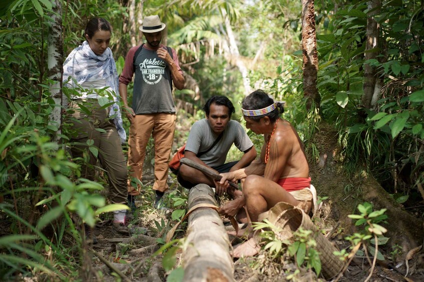 A group of people in a forested area, one person cutting a log with a machete. 3