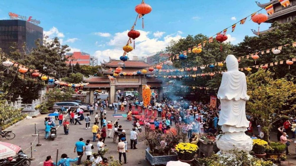 A crowded outdoor event with a large white statue, colorful lanterns, and a traditional building in the background. 2