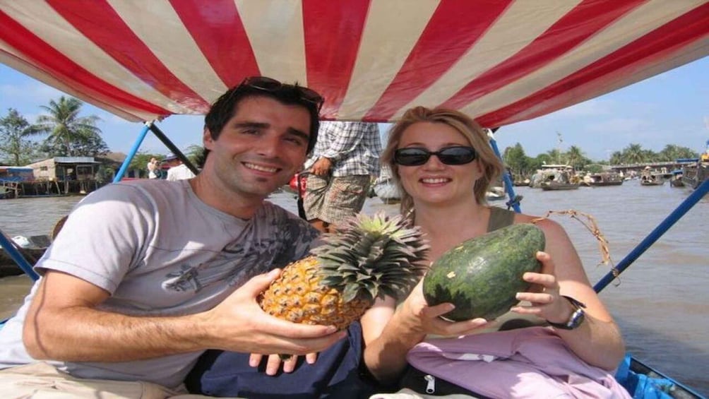 Two people holding a pineapple and a watermelon on a boat with a red and white striped canopy. 5