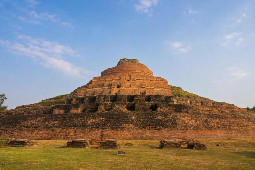 A large, ancient brick structure with a dome and multiple levels, surrounded by a grassy field. 1