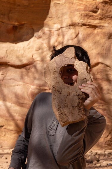 A person holding a rock with a hole in it, standing in front of a rocky cliff. 2