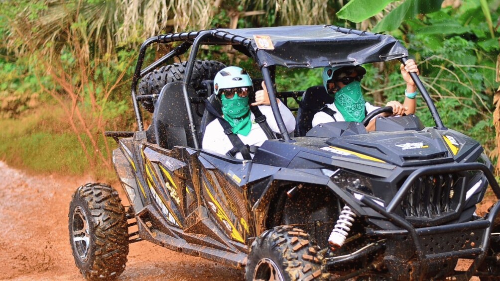 A four-wheeled off-road vehicle with a raised roof, muddy tires, and a driver wearing a helmet and a green bandana. 3