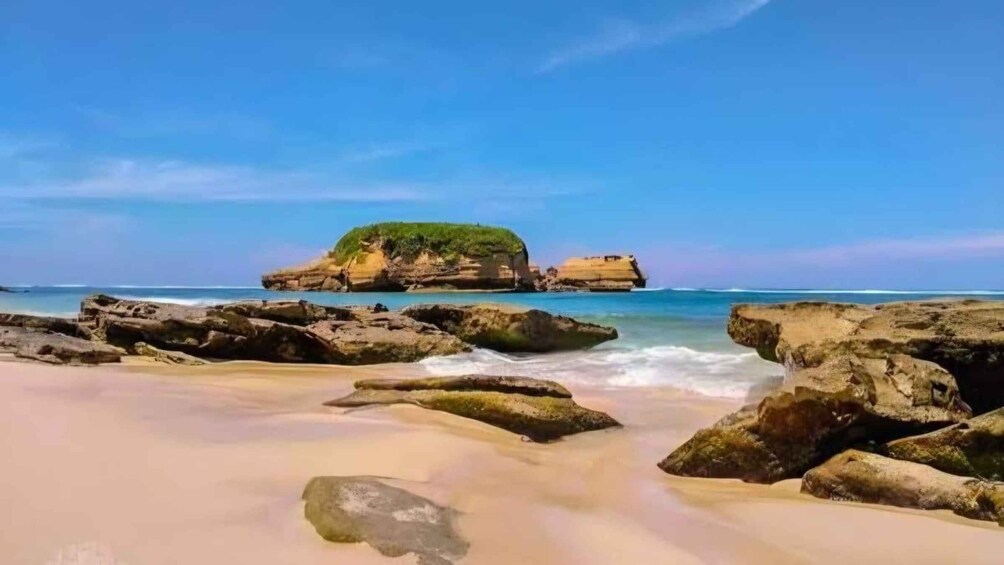 A beach with large rocks, clear blue water, and a small island with vegetation. 4