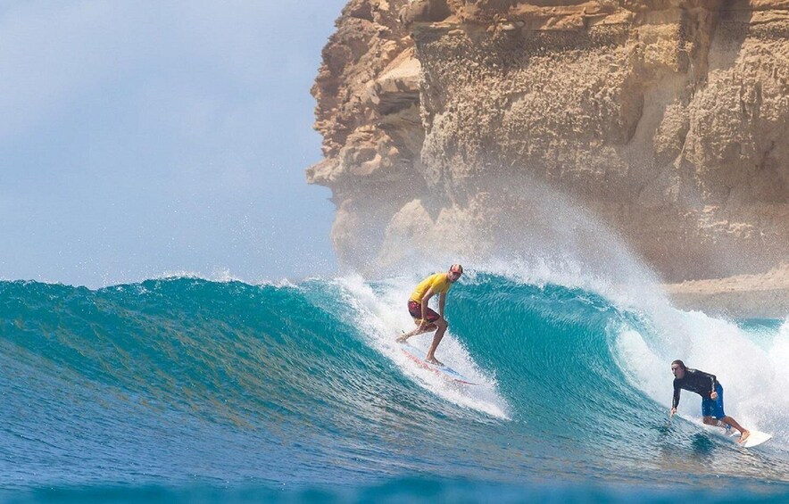 Two surfers riding a large wave with a rocky cliff in the background. 2