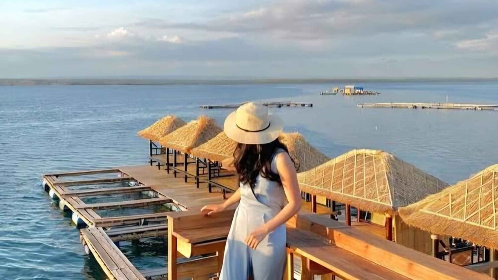 A woman on a wooden deck overlooking a calm sea with thatched-roof huts and a distant pier. 5