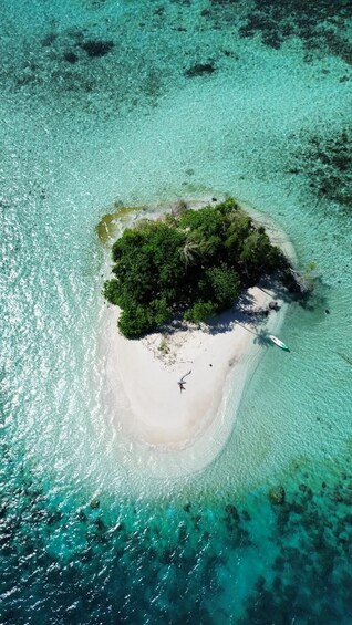 An aerial view of a small island with clear turquoise waters, a sandy beach, dense green vegetation, and a small boat anchored near the shore. 2
