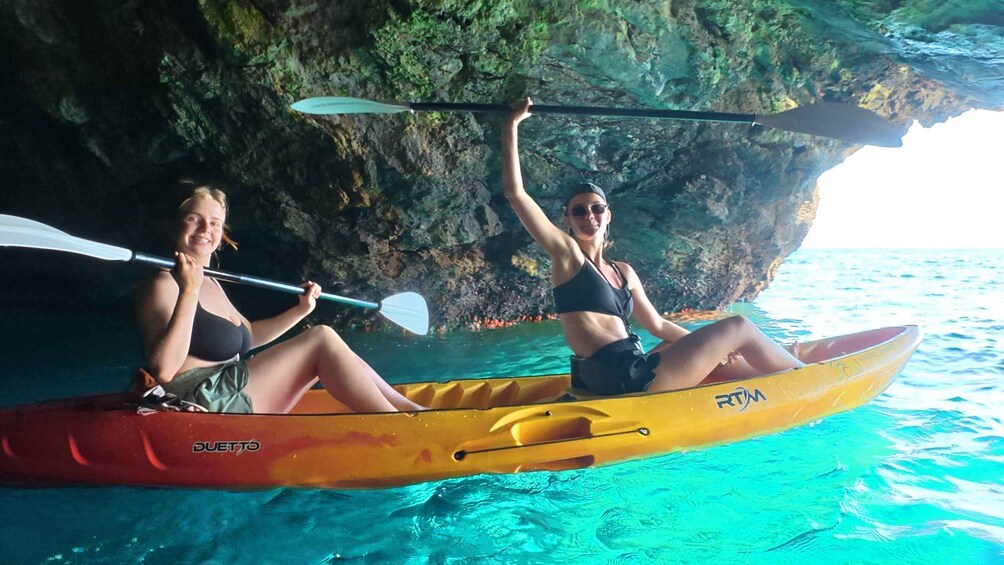 Two people kayaking in a yellow and red kayak with 'RTM' on it, surrounded by clear blue water and rocky cliffs. 2