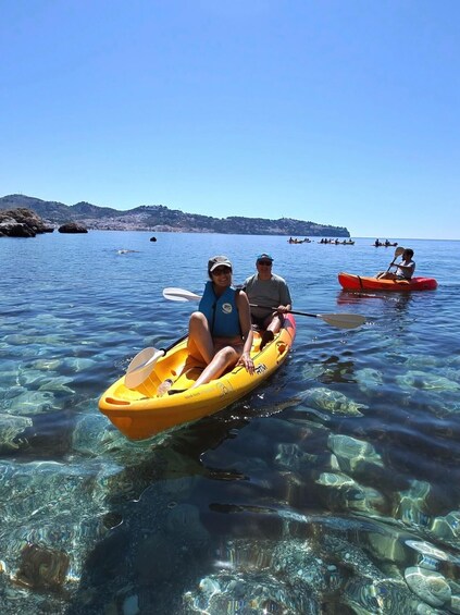 Two people kayaking on clear water with a rocky shoreline and a distant hill in the background. 5