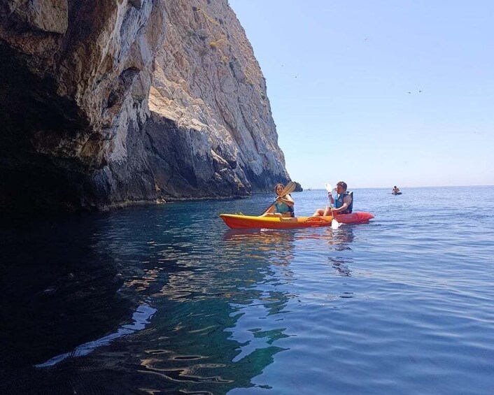Two people kayaking on calm sea waters near a rocky cliff. 1