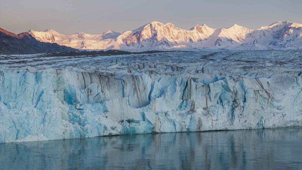 A glacier with a calm water surface reflecting the ice, and snow-capped mountains in the background. 2