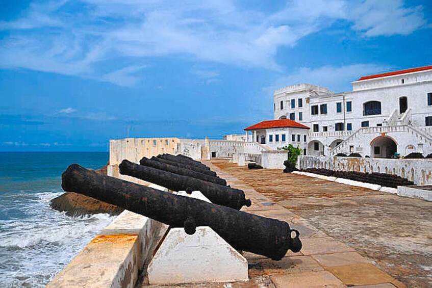 A coastal fortification with cannons, a stone wall, and a building with a red roof. 5