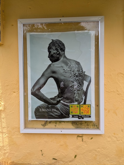 A framed black and white photograph of a seated man with the name 'EMANUEL' on it, mounted on a yellow wall. 1