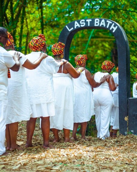 A group of people in white attire standing under a sign that reads 'LAST BATH' in a forested area. 1