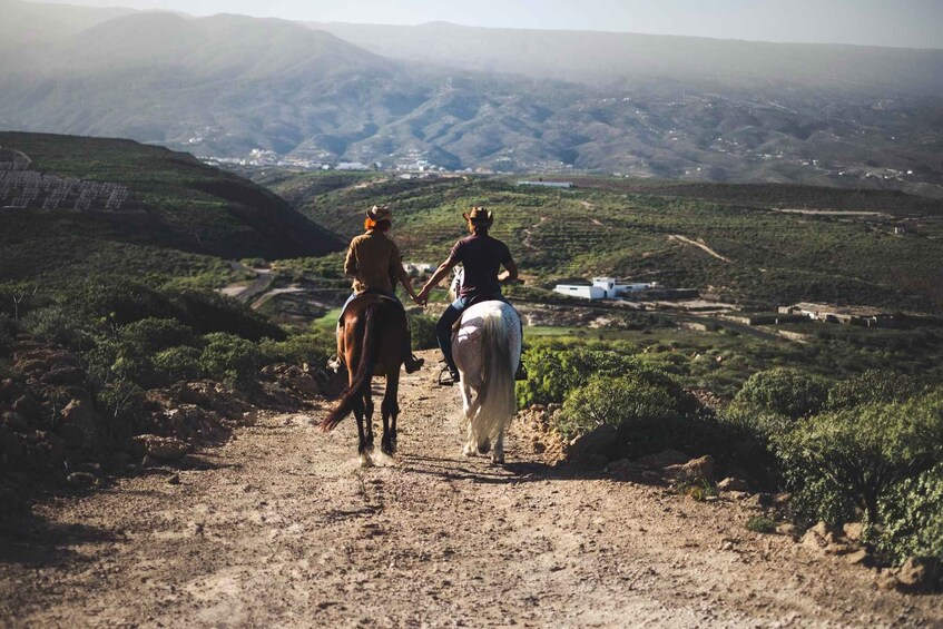 Two people horseback riding on a dirt path with a mountainous landscape in the background. 5