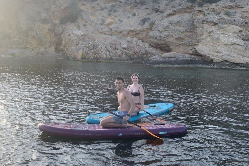 Two people on a paddleboard in calm water with rocky cliffs in the background. 3