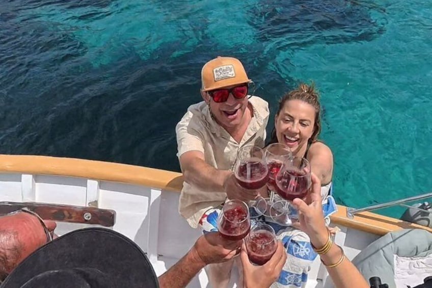 People on a boat, holding glasses of red liquid, with clear blue water in the background. 1